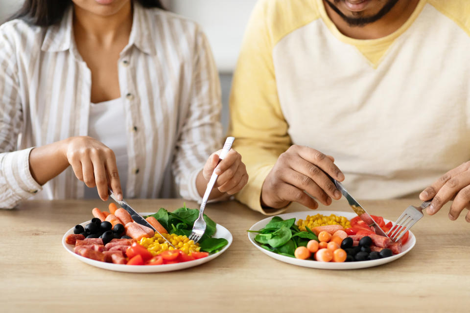 Two unidentified people dining on plates with assorted fresh vegetables, olives, corn and slices of meat