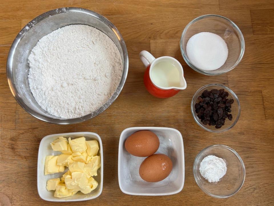 Ingredients for scones on a wooden table: flour, two eggs, butter, milk, sugar, sultanas, and baking powder.