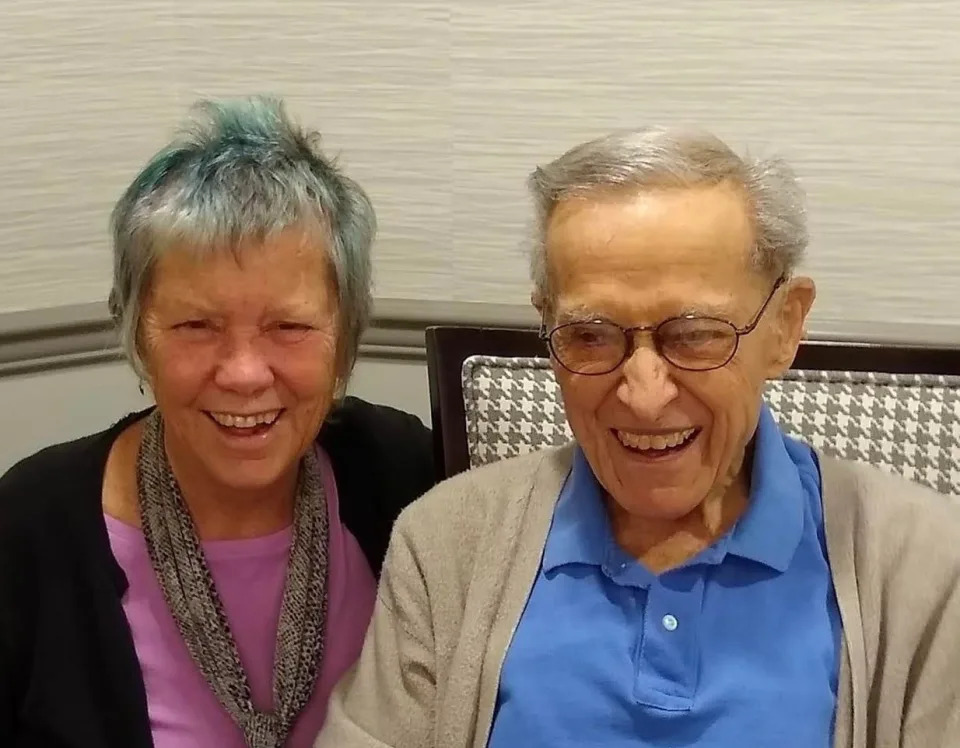 Louisa Rogers and her father sitting next to each other and smiling. Louisa has dyed blue short hair, dark eyes, and wears a black cardigan, pink t-shirt, and wears a scarf with a snakeskin print knotted around her neck. Her dad has white hair brushed to the left side, dark eyes, and wears dark-rimmed glasses. He looks off to the viewer's left side and wears a blue polo shirt and tan open sweater.