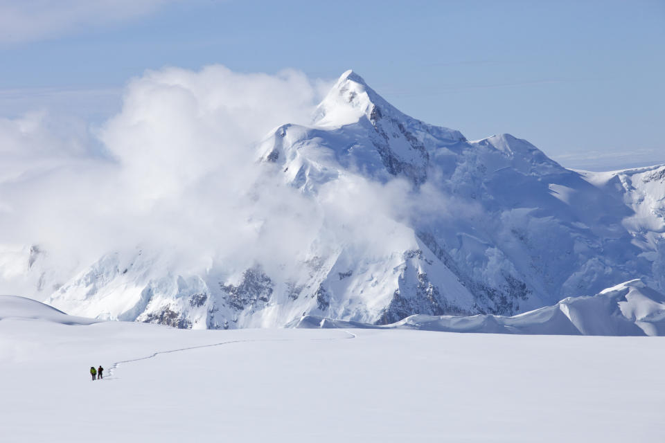 Two hikers trek across a vast snowy landscape with a large mountain in the background partially obscured by clouds