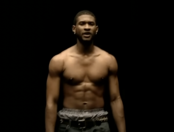 Usher shirtless, wearing dark pants, with a black background in a scene from a music video