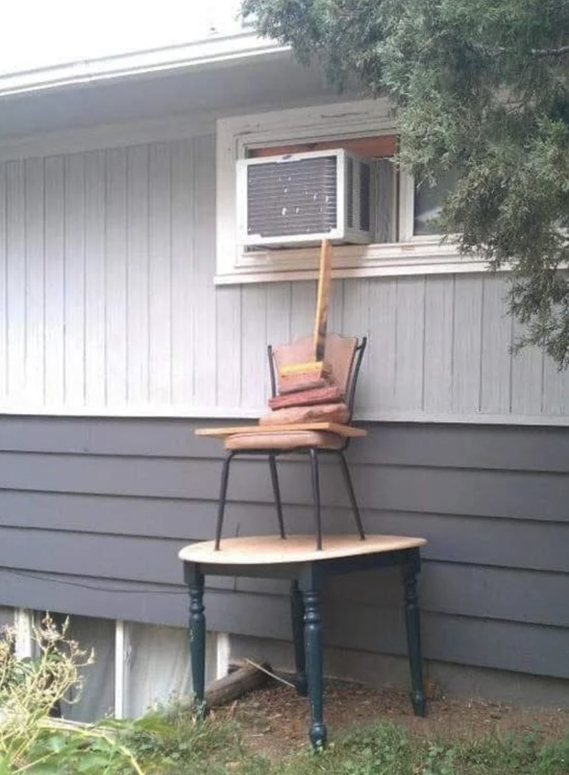 Window AC unit being propped up by a table, chair, and board