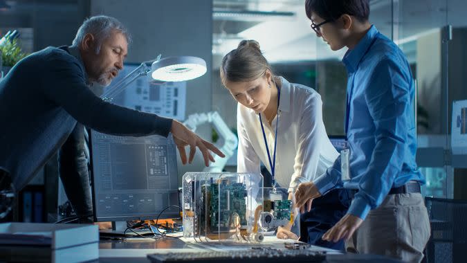 Team of Computer Engineers Lean on the Desk and Choose Printed Circuit Boards to Work with, Computer Shows Programming in Progress.