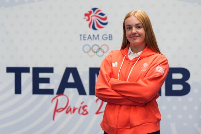 Archer Megan Havers, wearing an orange Team GB sweatshirt, poses with her arms crossed against a white wall featuring the team logo