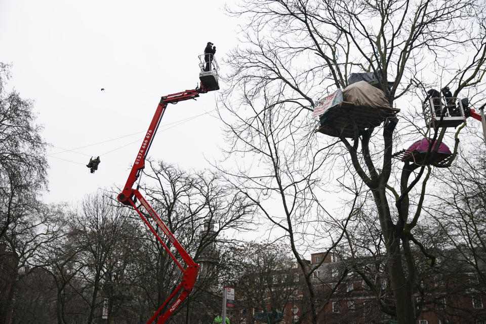 Two cherry pickers approach an HS2 Rebellion treetop camp, as a protester, left, uses a zip line between two trees, in an encampment in Euston Square Gardens in central London, Wednesday Jan. 27, 2021. Protesters against a high-speed rail link between London and the north of England said Wednesday that some of them have been evicted from a park in the capital after they dug tunnels and set up a makeshift camp. (Aaron Chown/PA via AP)