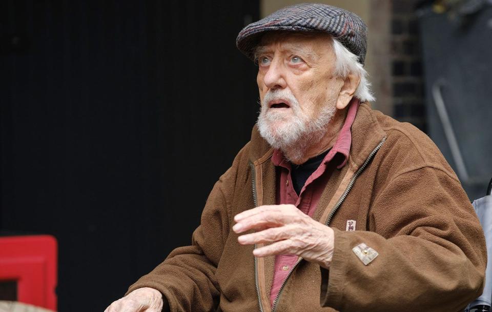 bernard cribbins in doctor who, an older man sits in a wheelchair looking up at someone off camera, he has grey hair and beard and wears a flat cap with brown coat