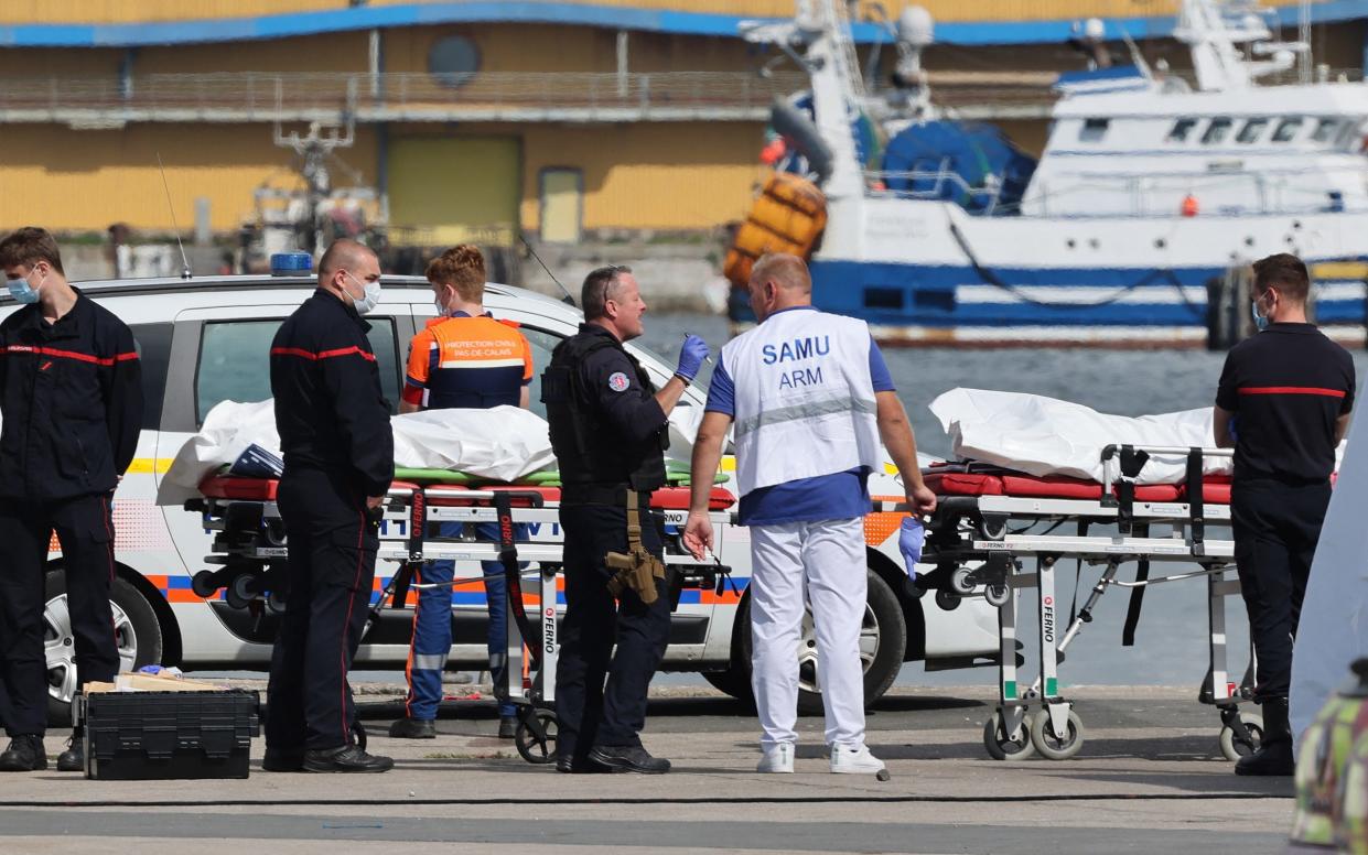 Emergency workers surround the two bodies, which are in white bodybags on wheeled trolleys, with moored boats seen in the background