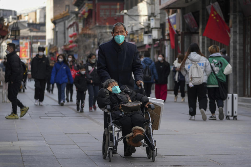An elderly man wearing a face mask pushes his partner in a wheelchair as visitors tour a pedestrian shopping street at Qianmen, in Beijing, Tuesday, Jan. 3, 2023. As the virus continues to rip through China, global organizations and governments have called on the country start sharing data while others have criticized its current numbers as meaningless. (AP Photo/Andy Wong)