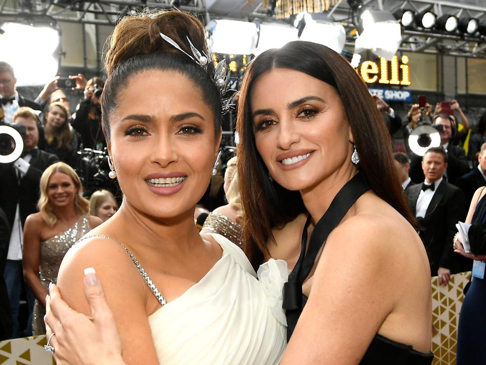 Salma smiling in a white gown with her arms around Penelope's waist and Penelope smiling in a black gown with her arms around Salma's torso.