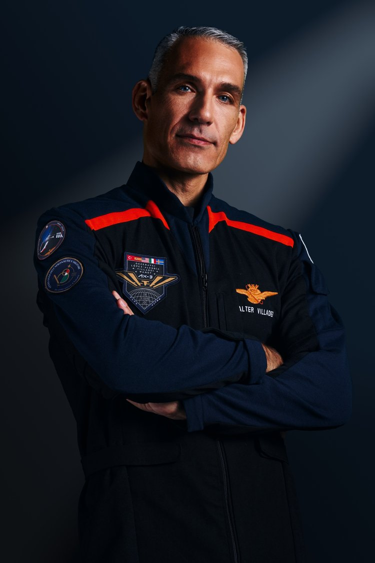 A man stands with arms crossed, wearing dark blue jumpsuits with red accented shoulder wings. He looks serious.