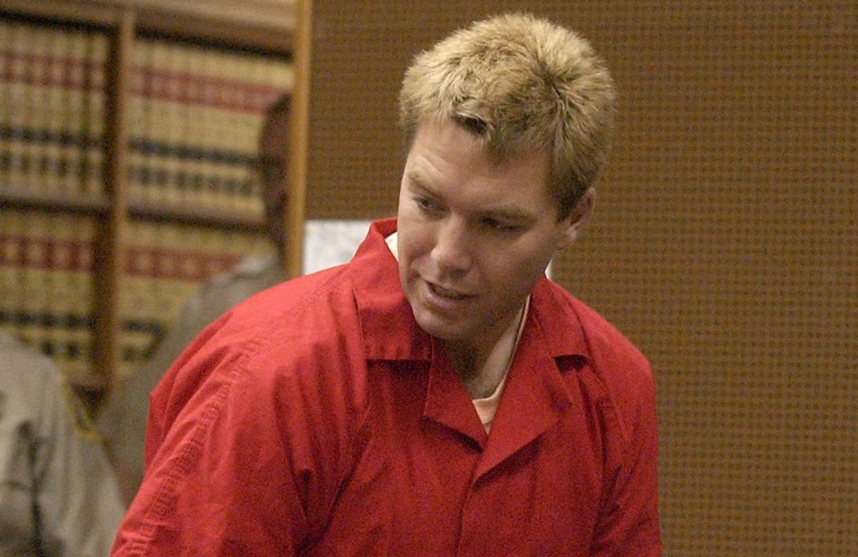 A man with blond hair wearing a red jumpsuit, looking to the left. Two police officers in brown shirts are stood behind him.