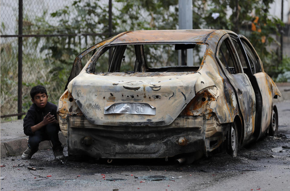 A boy takes cover behind a burned car after clashes erupt between protesters and riot policemen during a protest against deteriorating living conditions and strict coronavirus lockdown measures, in Tripoli, north Lebanon, Thursday, Jan. 28, 2021. Violent confrontations for three straight days between protesters and security forces in northern Lebanon left a 30-year-old man dead and more than 220 people injured, the state news agency said Thursday. (AP Photo/Hussein Malla)