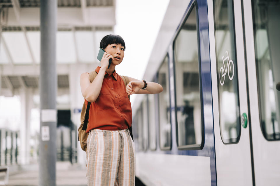 A person stands at a train station, talking on a cellphone while checking a watch. They carry a backpack and appear to be in a hurry