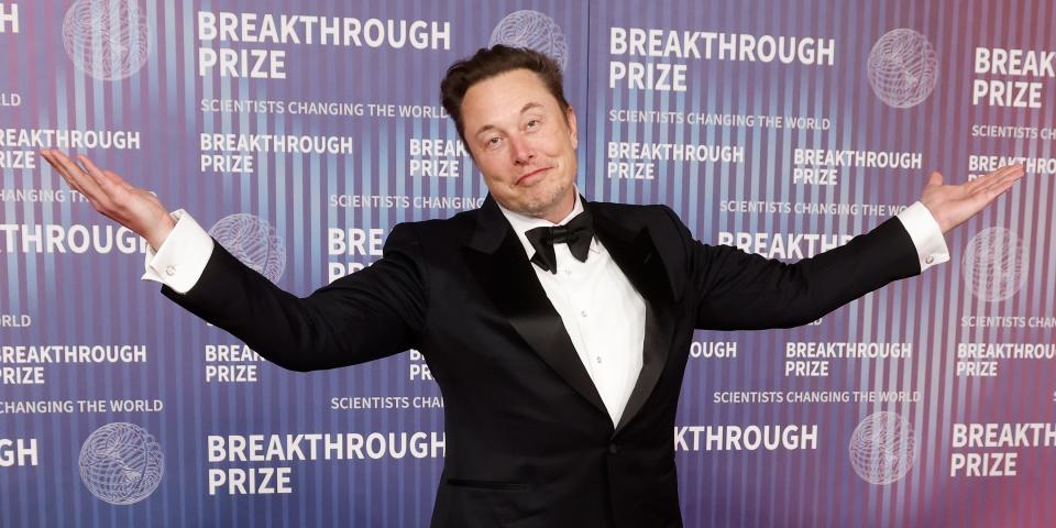 Elon Musk wearing a suit and bowtie whilst shrugging with his hands in the air