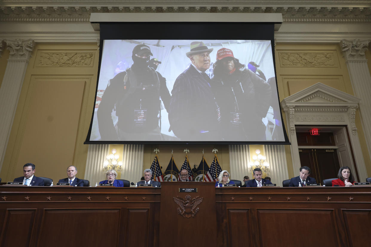 A screen placed above members of the committee shows Roger Stone in a fedora, with two people flanking him, one wearing a red MAGA baseball cap and one wearing a balaclava that hides most of his face and a dark baseball cap with Oath Keepers embroidered in yellow..