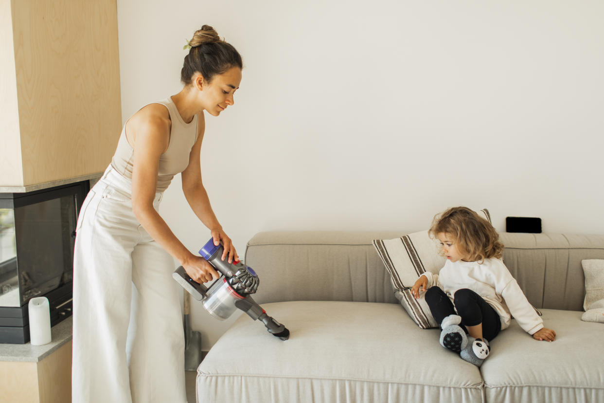 A young mother vacuums her sofa with a Dyson handheld vacuum while her young daughter watches