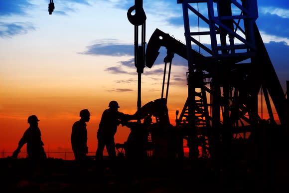 Men in hard hats near a gas rig in silhouette against a sky at sunset.