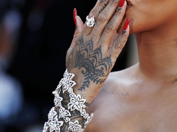 Rihanna blowing a kiss to fans on a red carpet while wearing small white sunglasses and a white dress