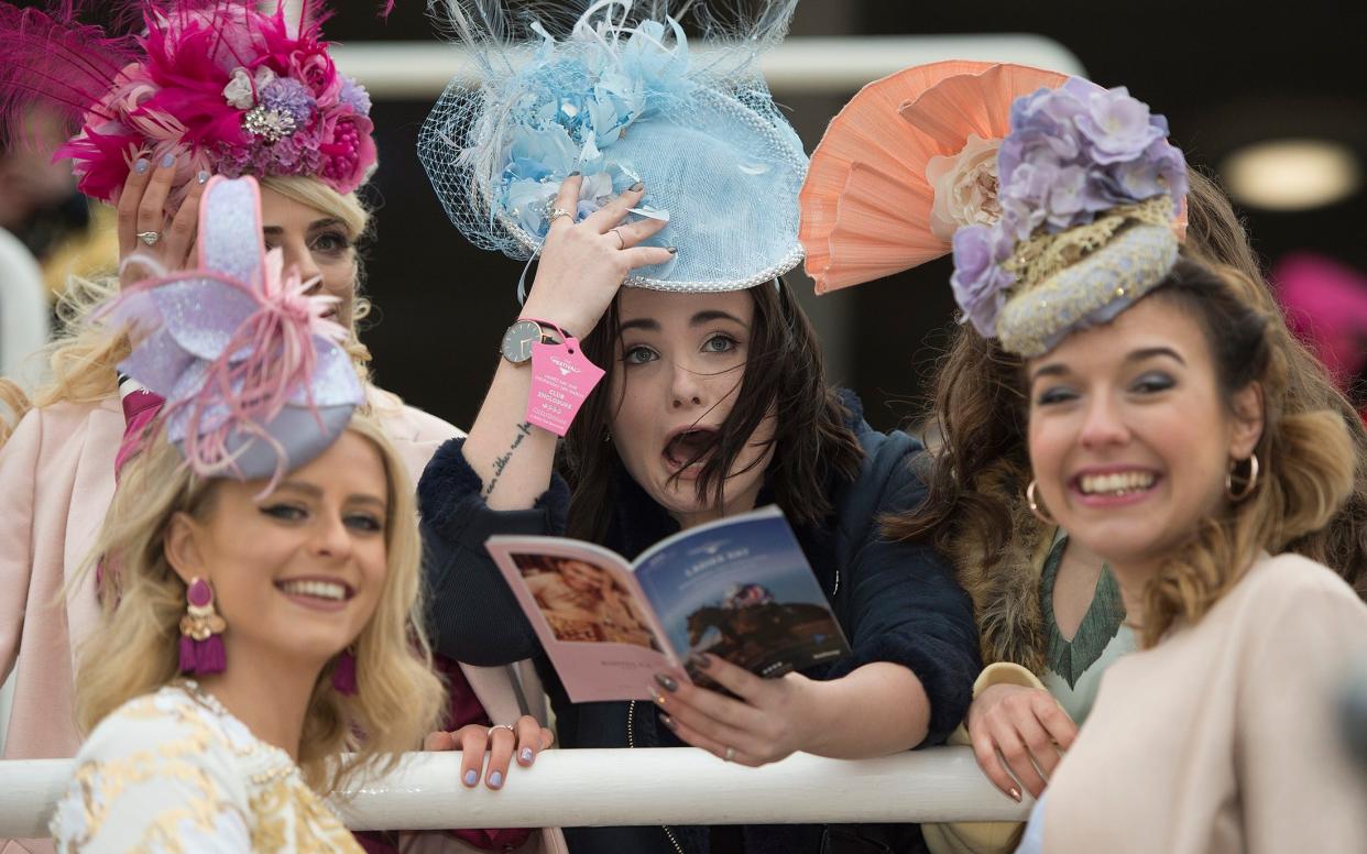 Three ladies in hats on Cheltenham Ladies' day, one reading a race program, holding onto her hat - ‘Outdated’ dress code scrapped for racegoers - The Telegraph/Eddie Mulholland