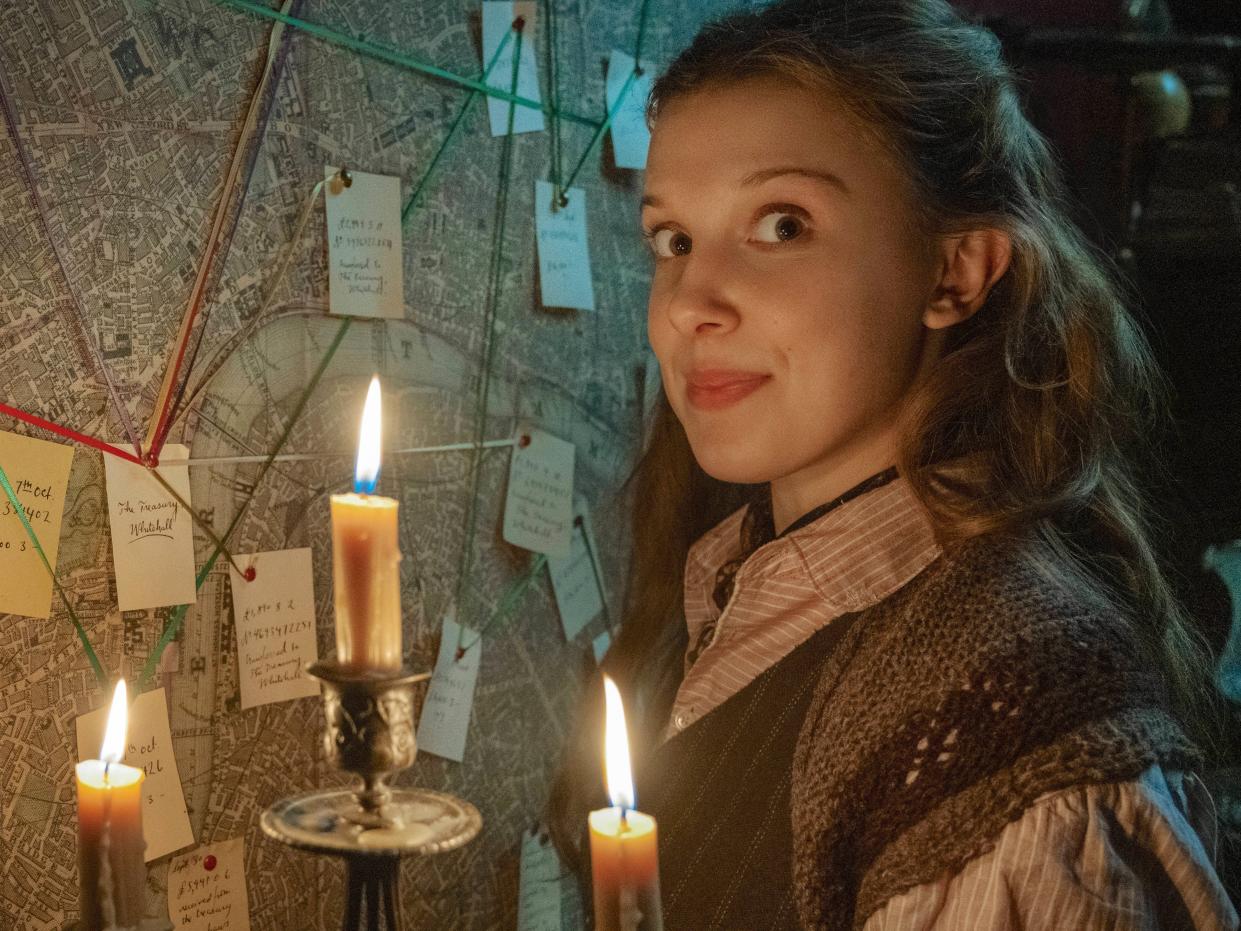 millie bobby brown as enola holmes, standing in front of a map with post-it notes and strings connecting them, illuminating the picture with a candelabra