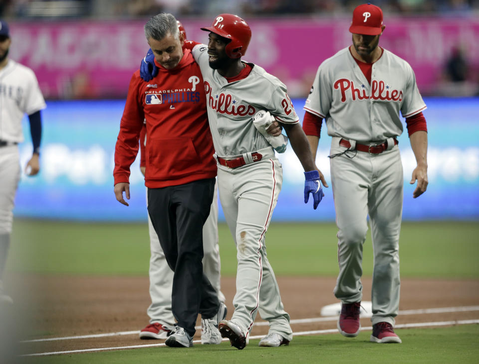 Philadelphia Phillies' Andrew McCutchen, center, is helped by a trainer after being injured while trying to get back to first base during the first inning of a baseball game against the San Diego Padres Monday, June 3, 2019, in San Diego. Phillies manager Gabe Kapler, right, looks on. (AP Photo/Gregory Bull)