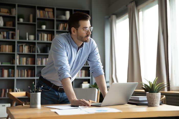 An investor stands in front of a laptop and looks pensively out a window.