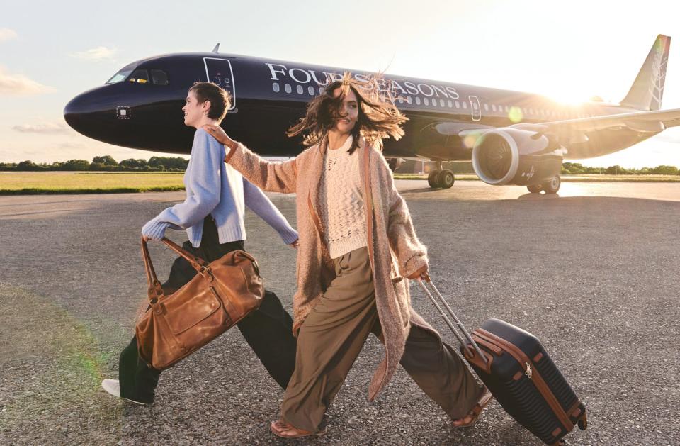 Two people walk with luggage on a windy day on the tarmac in front of Four Seasons' A321LR jet with a purple livery.