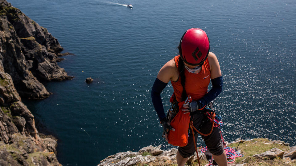 A woman stands on the edge of a cliff, water behind her, in climbing ropes and gear. She is adjusting a Sea to Summit Lightweight Dry Bag attached to her by a D-ring and karabiner.