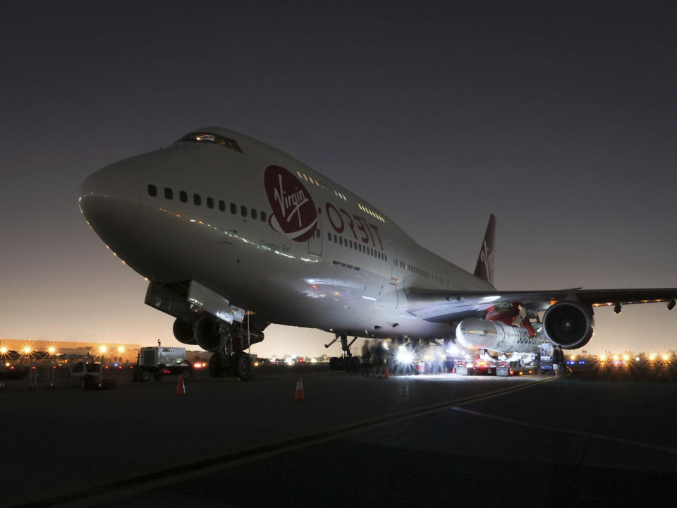 In this Wednesday, Oct. 24, 2018, photo released by Virgin Orbit, a completed LauncherOne rocket hangs from the wing of Cosmic Girl, a special Boeing 747 aircraft that is used as the rocket's "flying launch pad," at the Long Beach Airport in Long Beach, Calif. The system is intended to carry small satellites into orbit. Virgin Orbit is a sister company of Virgin Galactic, which is developing an air-launched rocket plane for carrying tourists on suborbital flights into space. (Greg Robinson/Virgin Orbit via AP)