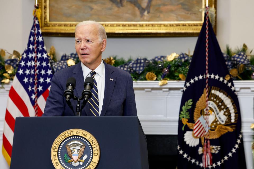 Biden speaking into microphones at a podium featuring the presidential seal, flanked behind him by the American flag and a flag showing the same seal. 