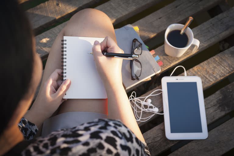 Young hipster woman writing journal on small notebook while sitting on wood bridge in morning time on weekend with high contrast sun lighting