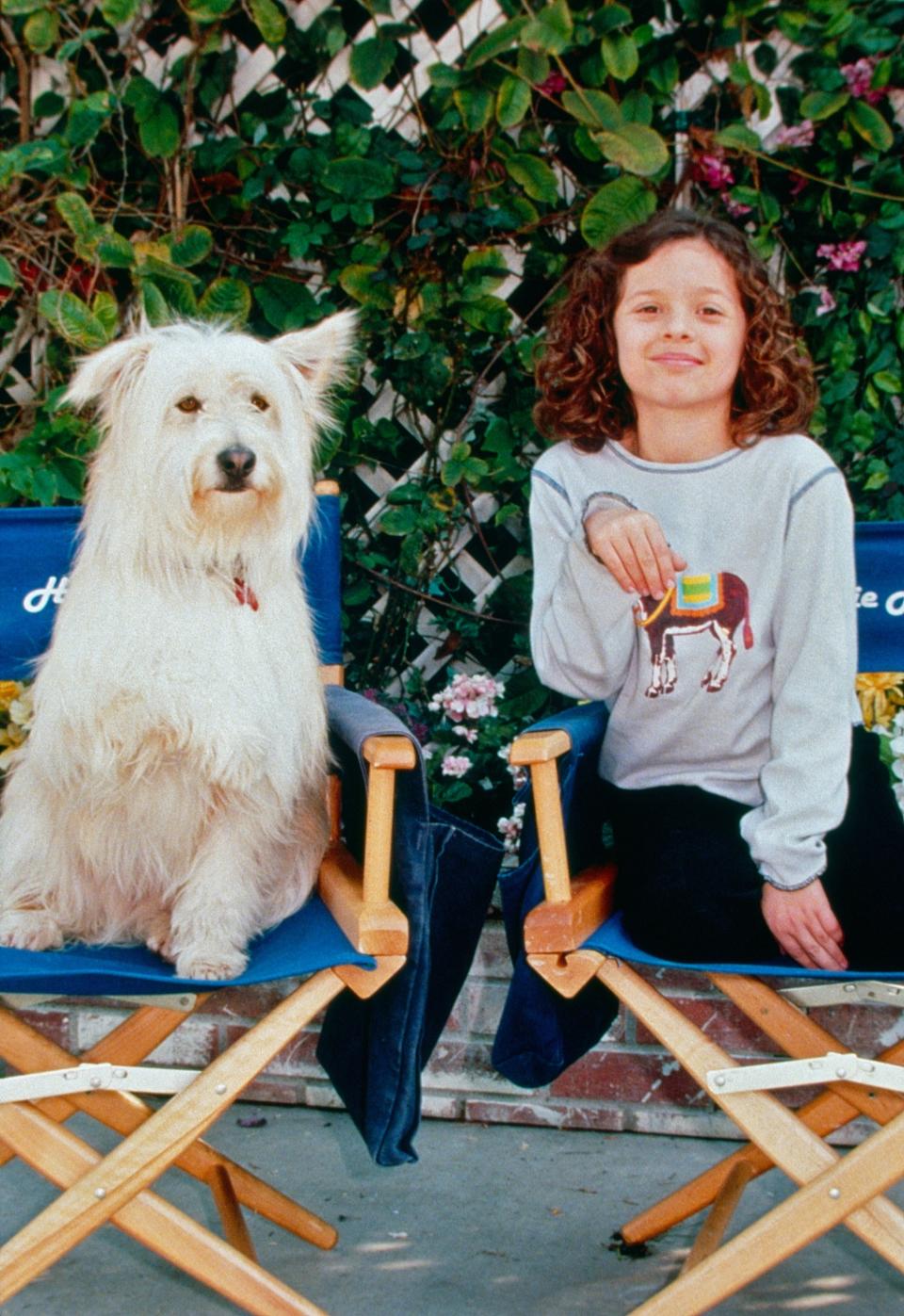 Child actor and a white fluffy dog sit on blue director's chairs in an outdoor setting with green foliage in the background