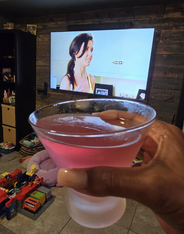 A hand holding a pink martini in front of a TV displaying a woman on "The Hills" show. Reddit comments are visible on the right