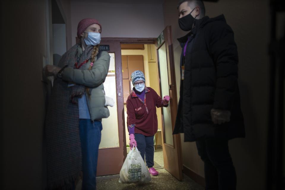Russian pensioner Galina Prokopieva, 72, wearing a face mask to protect against coronavirus, picks up a bag with products brought by the volunteers, also wearing face masks and observe social distancing guidelines wait for an elevator in Moscow, Russia, Sunday, Jan. 3, 2021. In the framework of Charitable action "New Year's Volunteers" of the "We Are Together" movement volunteers distribute food, medicine and other necessary things to elderly people. (AP Photo/Alexander Zemlianichenko)