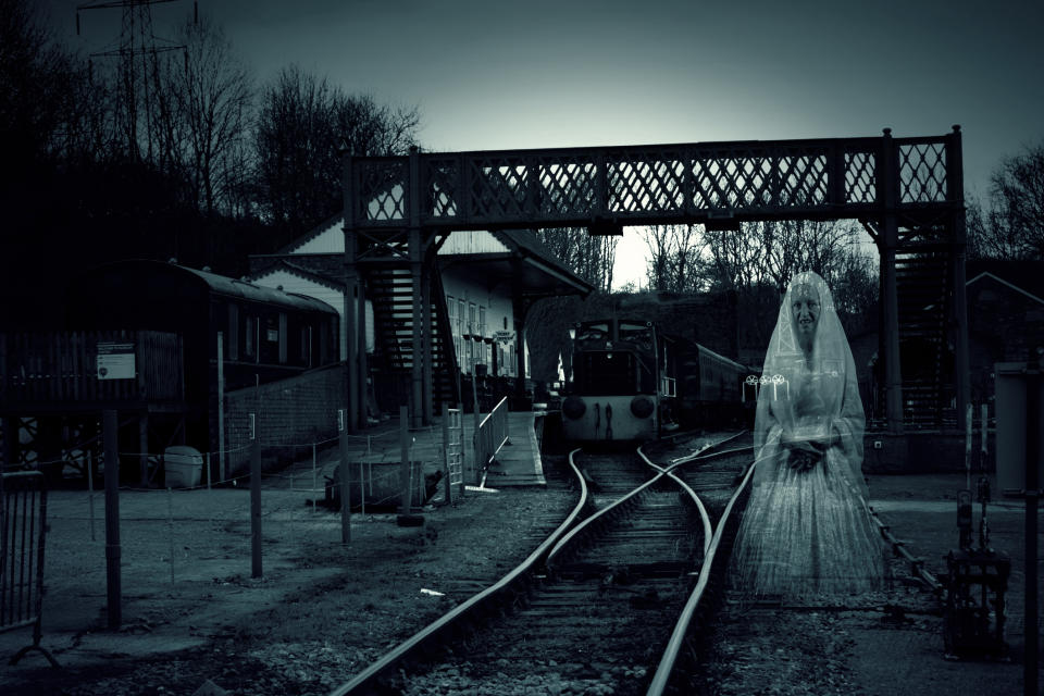 A ghostly figure in a wedding dress stands on a deserted railway track under a metal bridge. Surroundings are dark and eerie