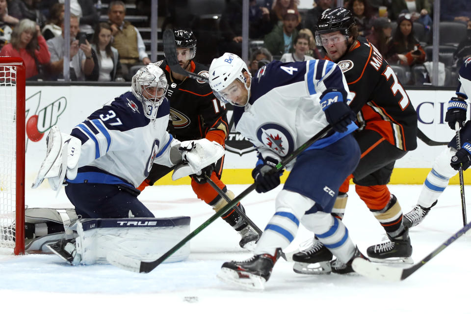 Winnipeg Jets defenseman Neal Pionk (4) clears the puck away from goaltender Connor Hellebuyck, left, with Anaheim Ducks centers Mason McTavish, right, and Adam Henrique (14) watching during the second period of an NHL hockey game in Anaheim, Calif., Wednesday, Oct. 13, 2021. (AP Photo/Alex Gallardo)