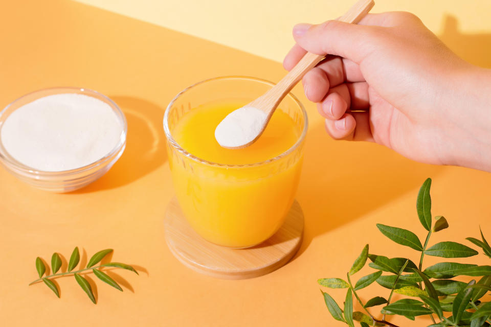 A hand is stirring a wooden spoon with white powder into a glass of orange juice