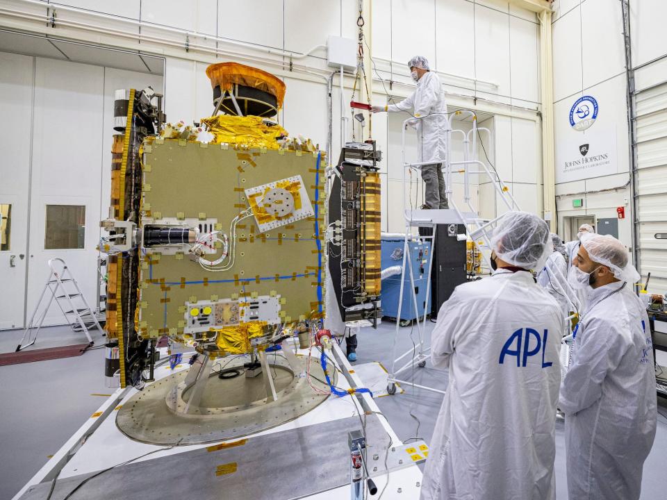 technicians in white cleansuits inspect dart spacecraft four foot yellow box in white warehouse cleanroom