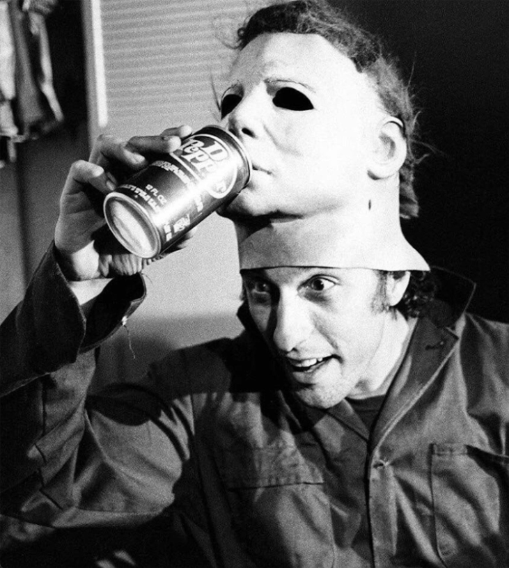 black and white photo of nick castle with a mask on top of his head, he's having the mask pretend to drink dr pepper