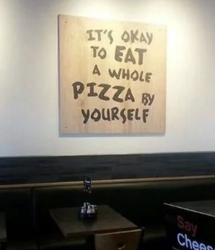 Wall sign reads, "It's okay to eat a whole pizza by yourself." Below the sign is a table with a small sign holder on it