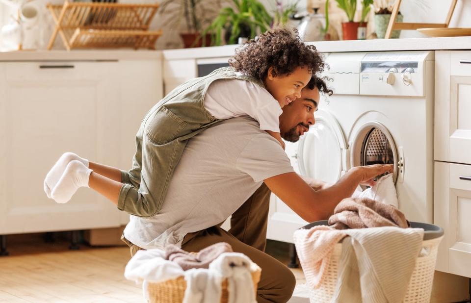 A child smiles while riding piggyback on an adult who is unloading clothes from a dryer. 