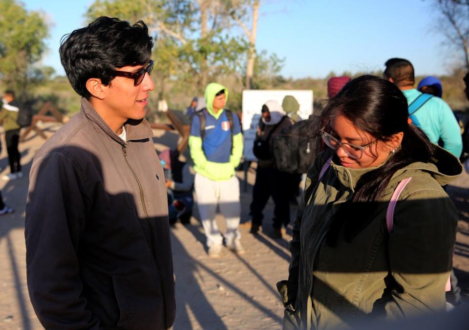 A young man and young woman standing in the partial shade and talking near a group of people