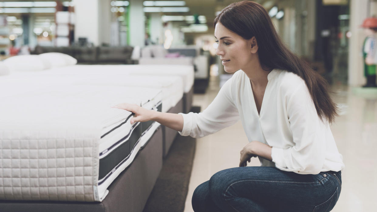  A woman with long dark hair looks at a mattress vs a mattress topper when shopping for a new bed. 