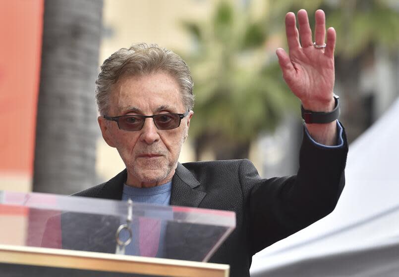 Frankie Valli in sunglasses, a blue shirt and a dark suit jacket holding up his left hand as he approaches a podium.