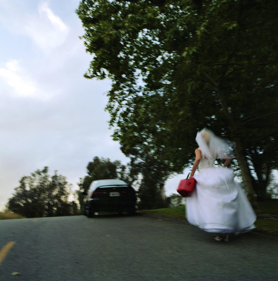 A bride, holding a red bag, walks along a road near parked cars and trees, possibly leaving her wedding ceremony