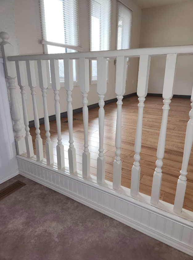 Image of a white spindle railing separating a carpeted area from a hardwood floor space with windows in the background