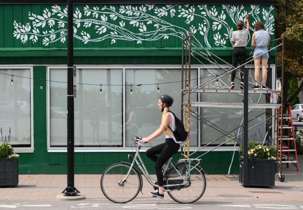 A cyclist rides past as Tori Lubbers and Emily Beckman, top right, paint a mural above the windows of the Green Door Restaurant in Ottawa, on Tuesday, Aug. 3, 2021.  (Justin Tang/The Canadian Press - image credit)