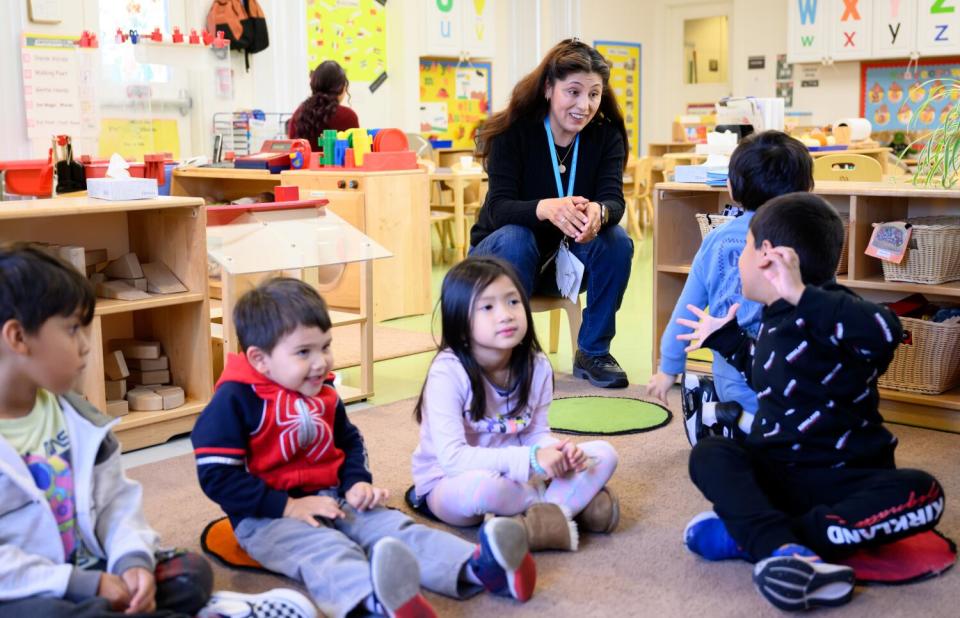 A preschool staff member watches children sitting on the classroom rug.