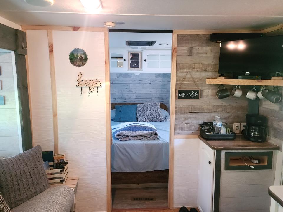 A small sitting area inside the RV, that includes a coffee maker, mugs, a TV, decorative signs, a cabinet, and a doorway to a bedroom.