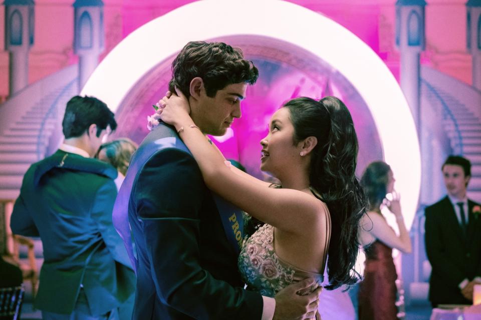 A teenage couple slow dances at their prom. Tall with dark brown hair, the boy wears a Prom King sash. The girl, with long black hair, looks at him smiling. He wears a suit. She wears a pink dress with embroidered flowers.